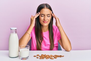 Young hispanic woman drinking healthy almond milk sitting on the table with hand on head, headache...