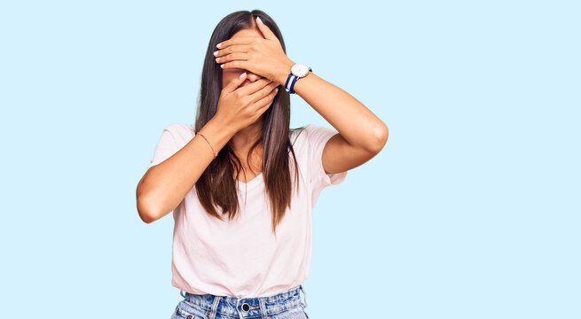 Young hispanic woman wearing casual white tshirt covering eyes and mouth with hands, surprised and shocked. hiding emotion