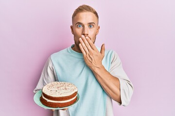 Young caucasian man chef cooking carrot cake covering mouth with hand, shocked and afraid for...