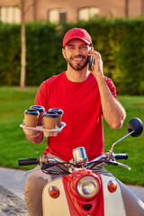 Young caucasian male courier talking by smartphone and smiling while delivering four coffee cups by scooter, standing on sunny street.