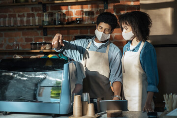 Young african american guy in apron and protective mask teaches trainee woman to work with espresso machine