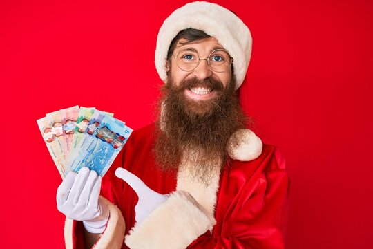 Handsome young red head man with long beard wearing santa claus costume holding canadian dollars smiling happy pointing with hand and finger