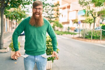 Young irish man with redhead beard smiling happy and showing his empty pockets jeans at the city.