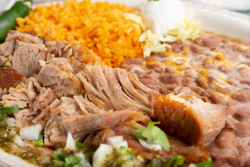 A closeup view of a plate of carnitas, with rice and beans sides, in a restaurant or kitchen setting.