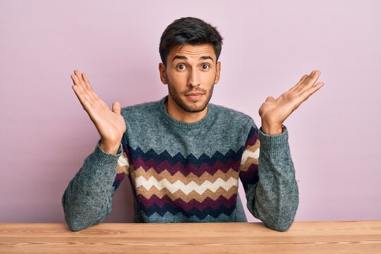 Young handsome man wearing casual winter sweater sitting on the table clueless and confused with open arms, no idea and doubtful face.