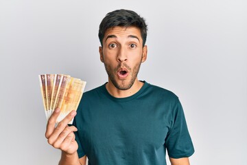 Young handsome man holding 500 norwegian krone banknotes scared and amazed with open mouth for...