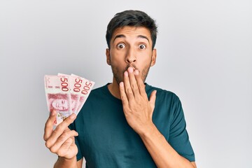 Young handsome man holding 500 swedish krona banknotes covering mouth with hand, shocked and afraid...
