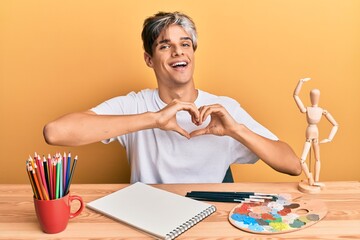 Young hispanic man artist sitting at the studio smiling in love doing heart symbol shape with hands. romantic concept.