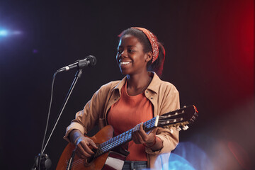 Waist up portrait of young African-American woman playing guitar on stage and singing to microphone...