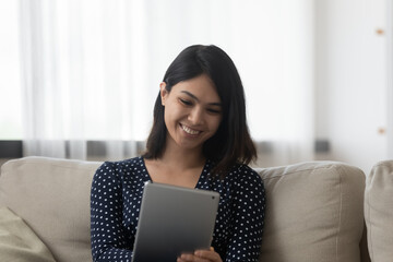 Close up smiling Asian young woman using tablet at home, sitting on cozy couch, happy attractive female looking at device screen, enjoying leisure time with gadget, chatting or watching video online