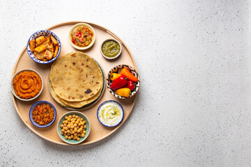 Indian Thali - selection of various dishes served on round wooden platter, white table. Assorted Indian vegetarian meze with chapati bread, paneer, dal, chutney in bowls. Top view, space for text