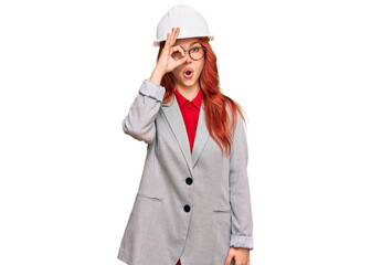 Young redhead woman wearing architect hardhat doing ok gesture shocked with surprised face, eye looking through fingers. unbelieving expression.