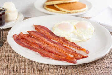 A view of a breakfast plate, featuring eggs over easy, bacon strips, and pancakes.