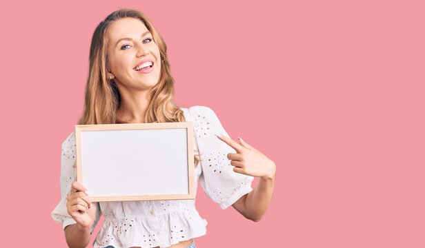 Young caucasian woman with blond hair holding blank empty banner smiling happy pointing with hand and finger