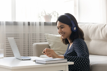 Smiling Asian young woman wearing headphones studying online at home, happy female student looking...