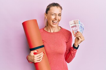 Beautiful caucasian sports woman holding yoga mat and swedish krona banknotes winking looking at...