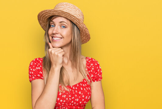 Beautiful caucasian woman wearing summer hat looking confident at the camera with smile with crossed arms and hand raised on chin. thinking positive.