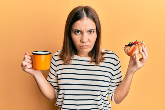 Young caucasian woman drinking coffee and eating pastry smiling looking to the side and staring away thinking.