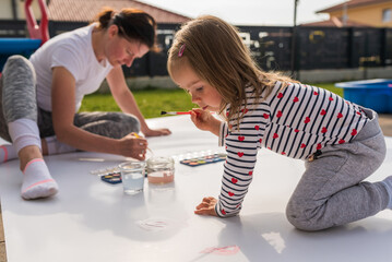 Child with mother painting with poster paint outdoors on the ground on big sheet of paper.