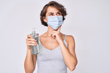 Young hispanic woman wearing medical mask holding hand sanitizer gel serious face thinking about question with hand on chin, thoughtful about confusing idea