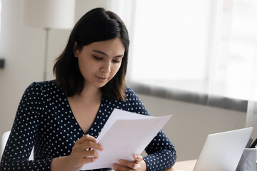 Close up serious Asian woman reading documents or letter, holding paper sheets, sitting at desk with laptop, confident businesswoman working with correspondence, research project, checking contract