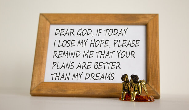 Wooden picture frame with Inspirational quote - 'Dear God, if today i lose my hope, please remind me that Your Plans are better than my dreams' on beautiful white fon. Two bronze angeles on the table.