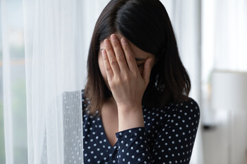 Close up frustrated Asian young woman covering face, crying, upset depressed female thinking about problems, standing near window, suffering from break up with boyfriend or divorce, health trouble