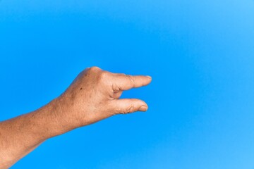 Senior man arm and hand over blue isolated background holding invisible object, empty hand doing clipping and grabbing gesture