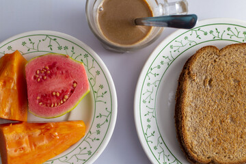 Café da manhã; Um prato com frutas, uma torrada e uma xícara de leite com café sobre...