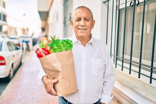 Senior man smiling happy holding paper bag with food walking at the city.