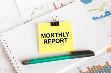 A yellow sticker with text Monthly Report is in a Notepad with a green pen financial charts and documents