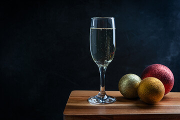 Wineglass full of dry white Prosecco sparkling champagne with golden and red christmas or new year decoration balls on dark brown wooden bar counter against black background. Image with copy space