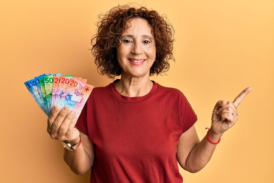 Beautiful middle age mature woman holding swiss franc banknotes smiling happy pointing with hand and finger to the side