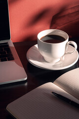 Laptop, cup of coffee, notepad and pen in the morning light with shadows. Home office desktop. Educational and business concept.