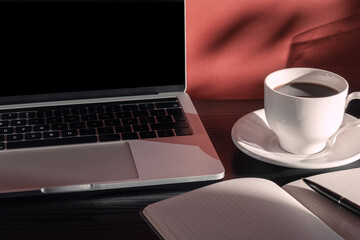 Laptop, cup of coffee, notepad and pen in the morning light with shadows. Home office desktop. Educational and business concept.