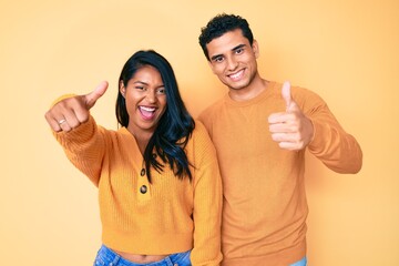 Beautiful latin young couple wearing casual clothes together approving doing positive gesture with hand, thumbs up smiling and happy for success. winner gesture.