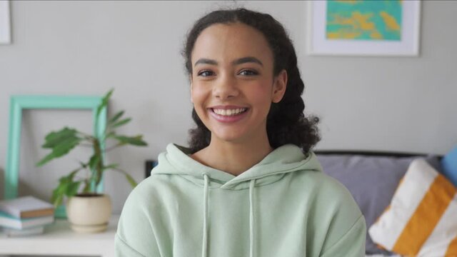 Happy african american teen girl looking at camera at home. Cheerful generation z mixed race pretty teenager smiling while sitting on bed in bedroom casual interior, headshot portrait.