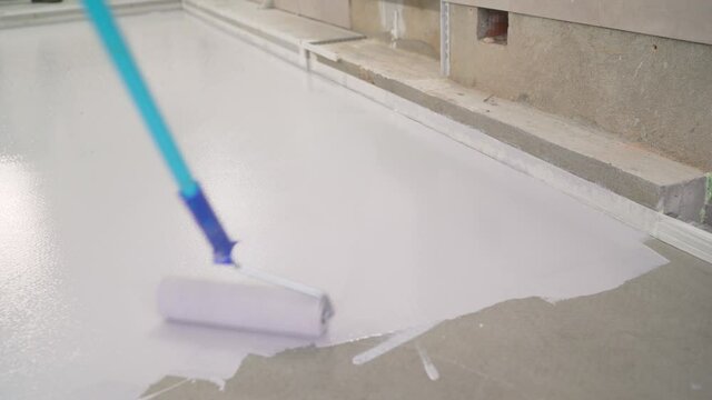 A worker paints the concrete floor with white paint. A worker paints the floor with a roller. Manual painting of a white floor with a paint roller for waterproofing