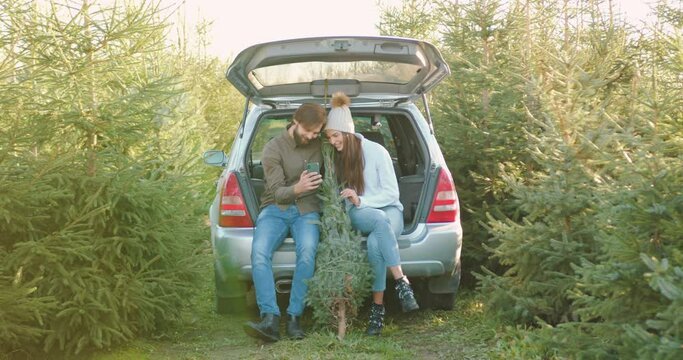 Pleasant lucky joyful young couple sitting in car's trunk among green fir trees and reviewing photos on smartphone ,close up ,slow motion