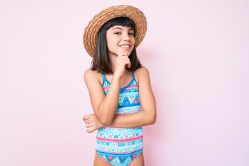 Young little girl with bang wearing swimsuit and summer hat smiling looking confident at the camera with crossed arms and hand on chin. thinking positive.