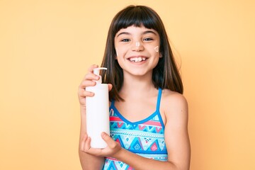 Young little girl with bang wearing swimsuit and holding sunscreen lotion looking positive and happy standing and smiling with a confident smile showing teeth