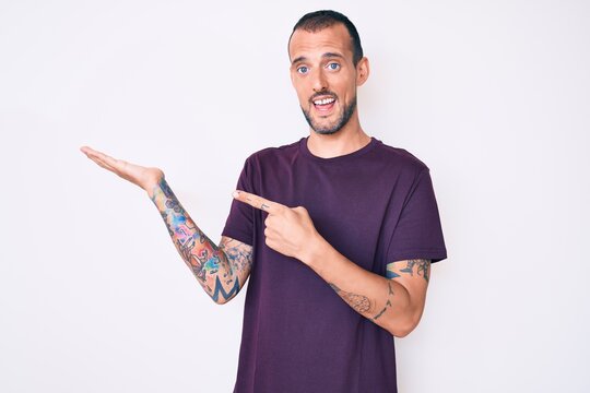 Young handsome man with tattoo wearing casual clothes amazed and smiling to the camera while presenting with hand and pointing with finger.