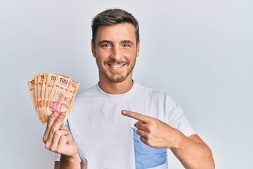 Handsome caucasian man holding mexican pesos smiling happy pointing with hand and finger