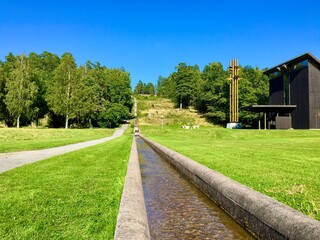 A canal with fresh, clear and cold water next to a small asphalt road. A wooden building of a chapel at the other side. Natural and great view of something man made. Jarfalla, Stockholm, Sweden.