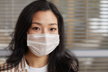 Close up portrait of young Asian woman wearing mask and looking at camera against office blinds background, copy space