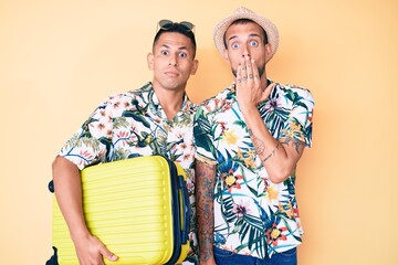 Young gay couple of two men holding suitcase going on summer vacation covering mouth with hand, shocked and afraid for mistake. surprised expression