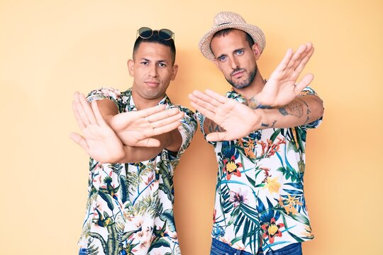 Young gay couple of two men wearing summer hat and hawaiian shirt rejection expression crossing arms and palms doing negative sign, angry face