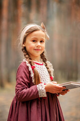 Little cute girl in a historical dress walks in a pine forest with a book in her hands