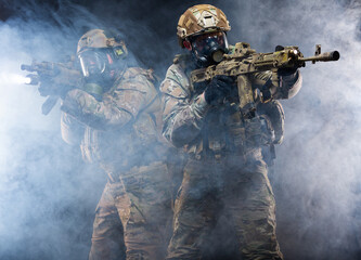 Two soldiers in military gear, bulletproof vests and gas masks cover each other and raise their submachine guns taking aim, in full combat readiness to break through the smoke from chemical weapons