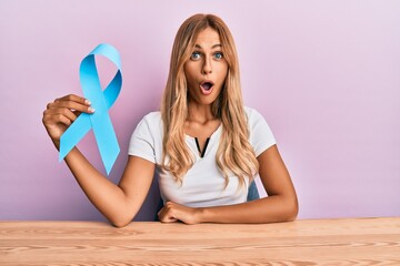 Beautiful blonde young woman holding blue ribbon scared and amazed with open mouth for surprise,...